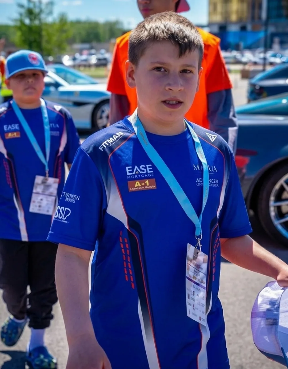 Young team member in a royal blue sublimated team tee with sponsor logos on the chest and sleeves, wearing an event lanyard in the paddock.