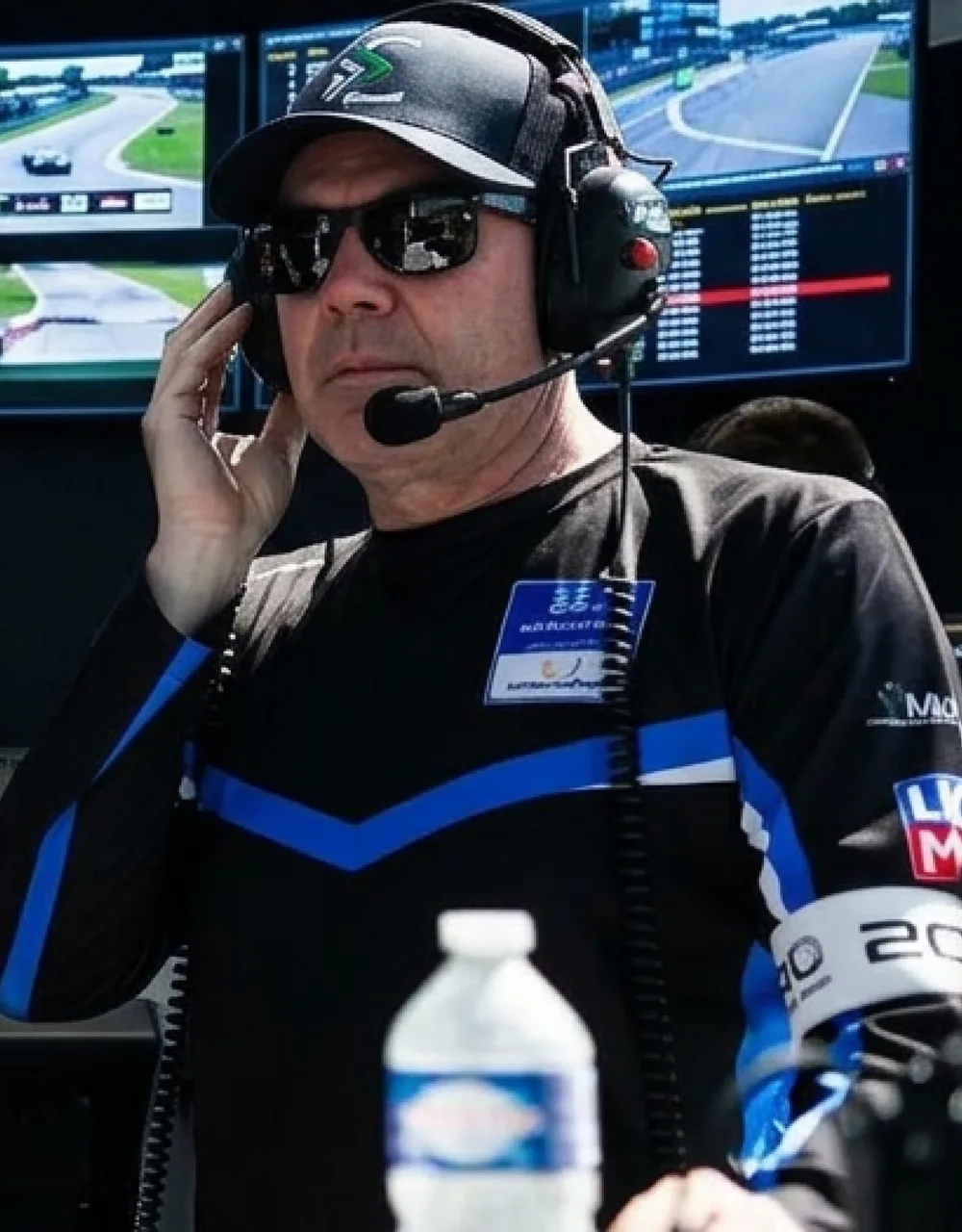 Crew member in a black sublimated team tee with a blue chevron across the chest, wearing a headset in front of the timing screens.